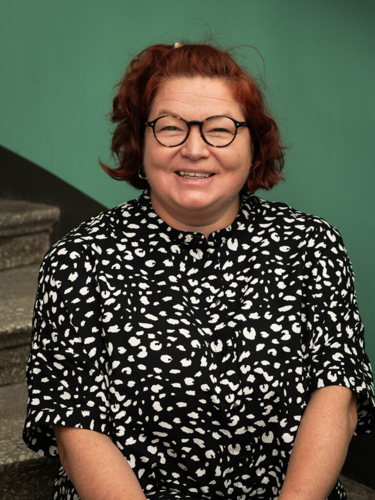 Kuvassa Kristiina Vesamalla on silmä lasit, punertava tukka ja musta paita jossa valkoisia pilkkuja. 