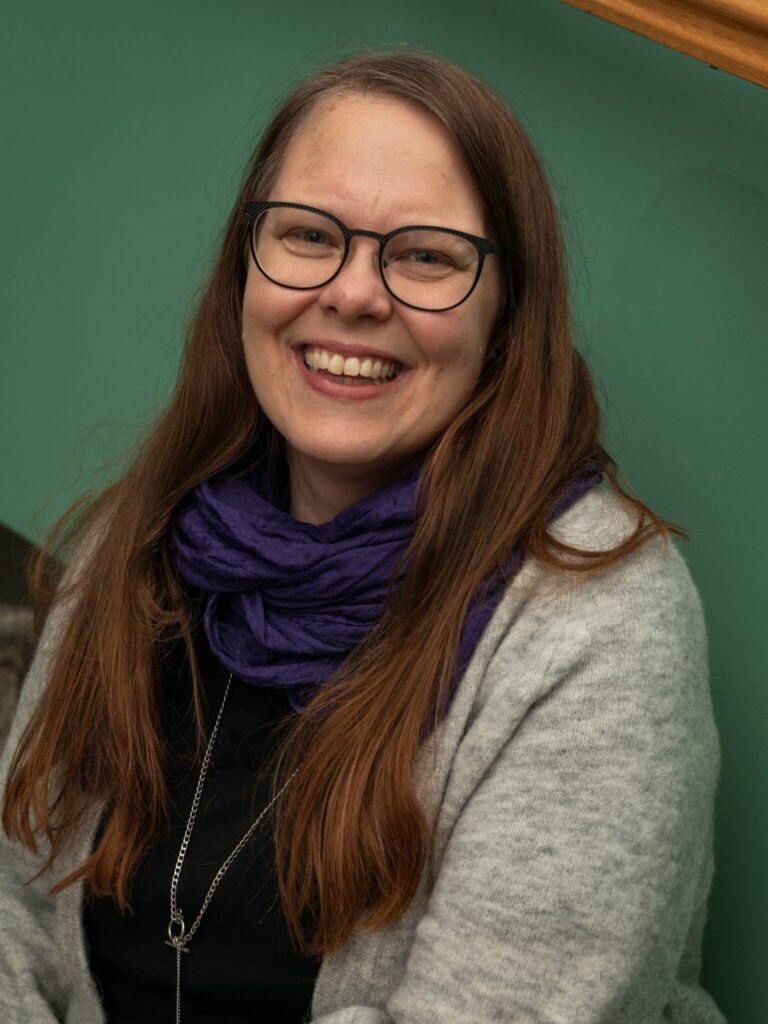 Heli Laurikaisella on pitkät punertavat hiukset ja tummat silmälasit sekä harmaa villatakki ja violetti huivi. 